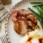 Bacon wrapped pork tenderloin on a plate cut into medallions with mashed potatoes and green beans next to it.