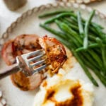 Bacon wrapped pork tenderloin on a plate cut into medallions with mashed potatoes and green beans next to it, and a fork taking one bite of the pork.