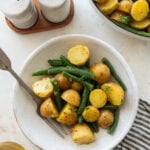 One serving of green beans and potatoes in a bowl with a fork, with the serving bowl and other ingredients nearby.