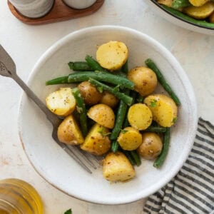One serving of green beans and potatoes in a bowl with a fork, with the serving bowl and other ingredients nearby.