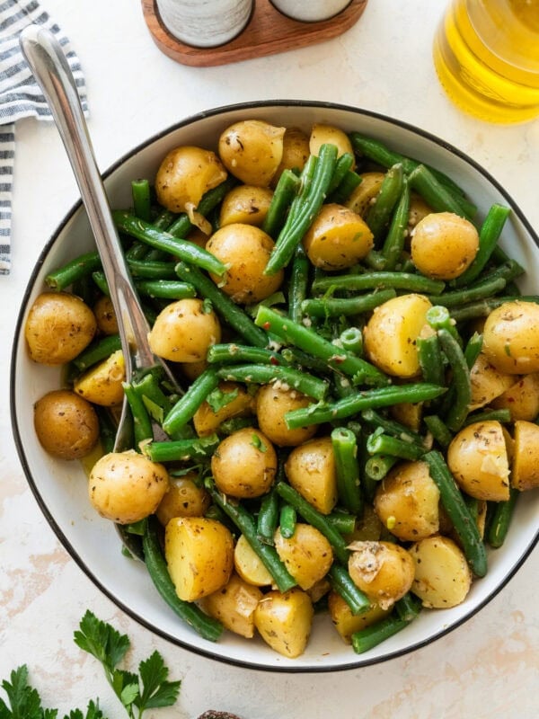 A large bowl of green beans and potatoes with more ingredients nearby and a utensil resting inside the bowl.