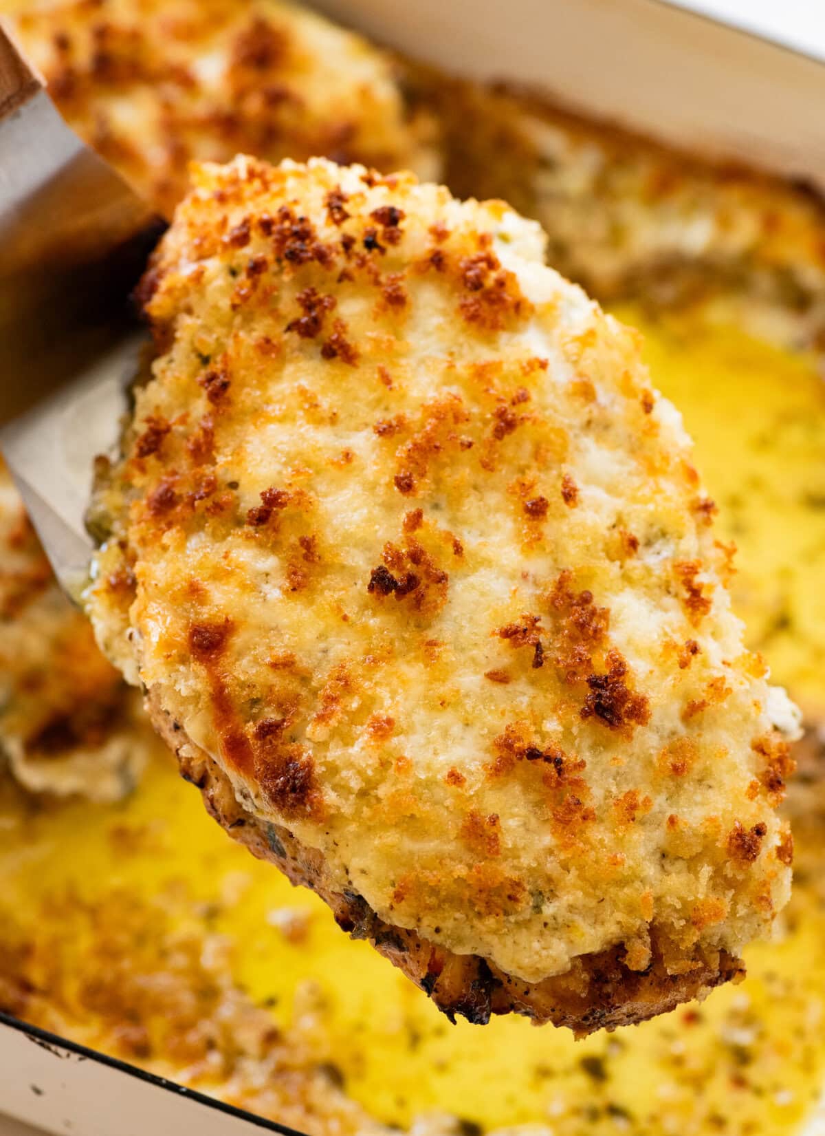a parmesan crusted chicken breast being held with a utensil over a pan after baking and broiling.