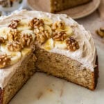 Frosted banana nut cake garnished with nuts and banana slices, with a slice removed.