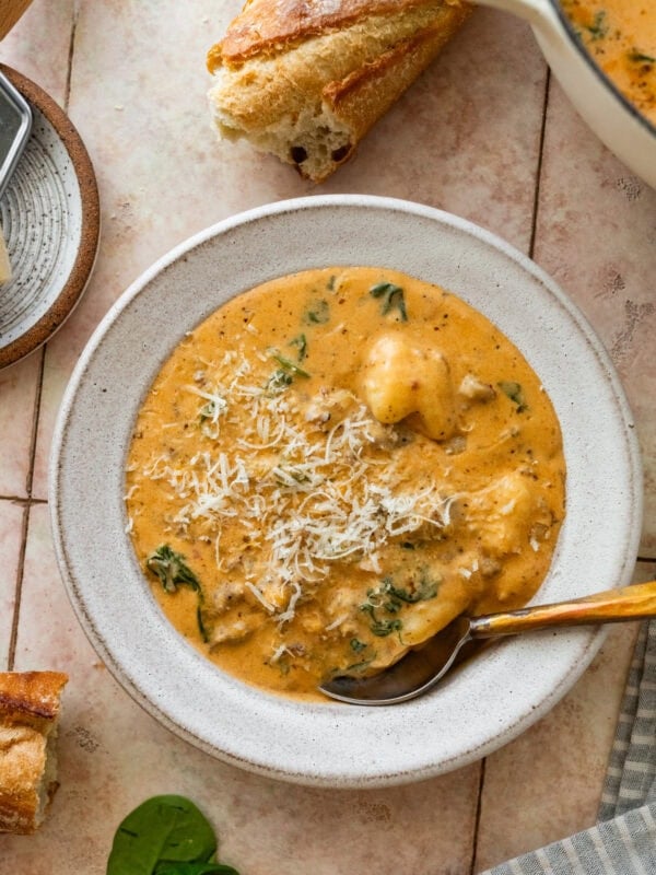 A bowl of sausage gnocchi soup with a spoon resting inside and more ingredients nearby.