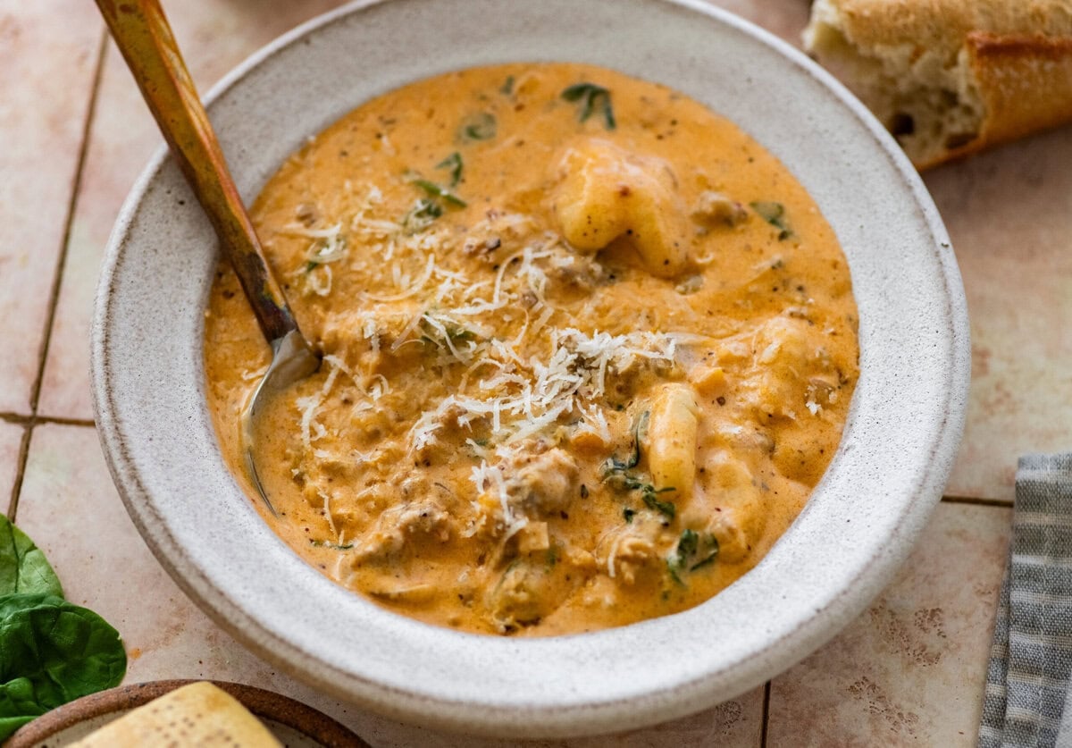 A bowl of sausage gnocchi soup with a spoon resting inside and more ingredients nearby.
