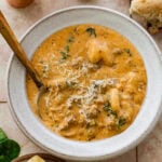 A bowl of sausage gnocchi soup with a spoon resting inside and more ingredients nearby.