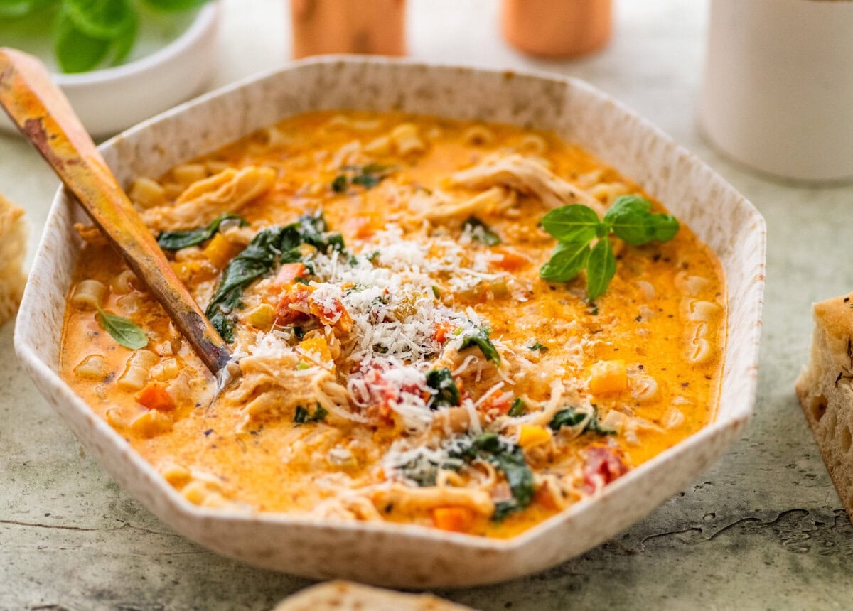 A bowl of tuscan chicken soup with more ingredients nearby and a spoon inside the bowl.