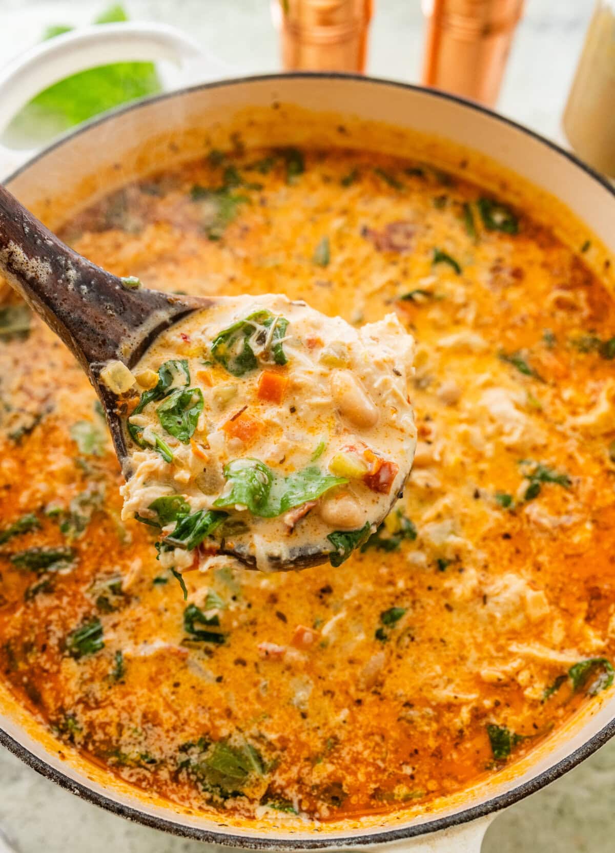 A pot of tuscan chicken soup with more ingredients nearby and a spoon holding some up.